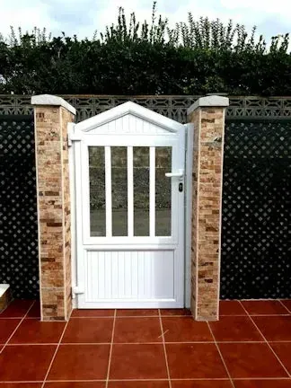 Puerta blanca con paneles verticales y barras horizontales, enmarcada por columnas de ladrillo, sobre un fondo verde oscuro.