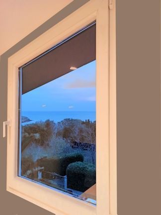 Ventana con vista a un cuerpo de agua y árboles bajo un cielo azul y morado, parcialmente cubierta por una persiana cerrada de color oscuro.