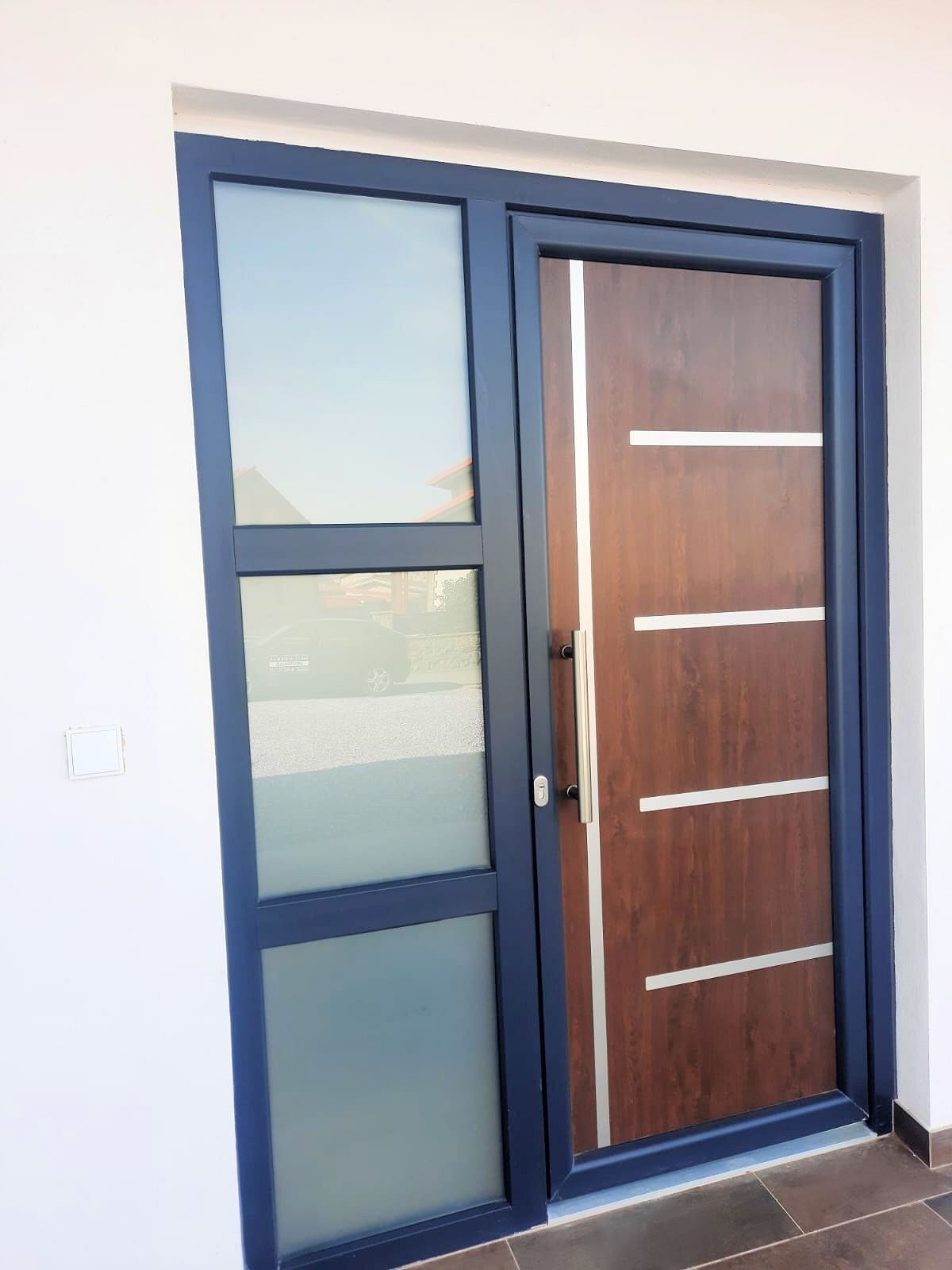 Puerta exterior con marco azul oscuro, panel de madera marrón y ventana lateral de vidrio. El tirador es vertical.