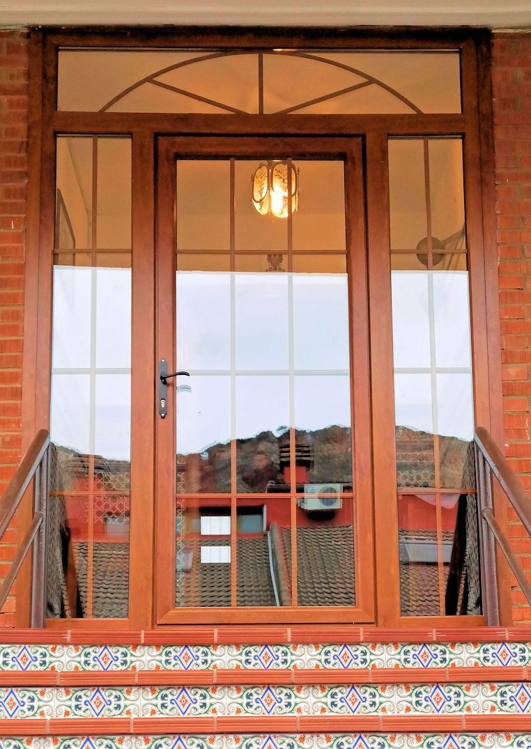 Puerta principal de madera con paneles de vidrio, ventanas laterales y travesaño. Refleja una vista de los tejados y una lámpara en el interior.