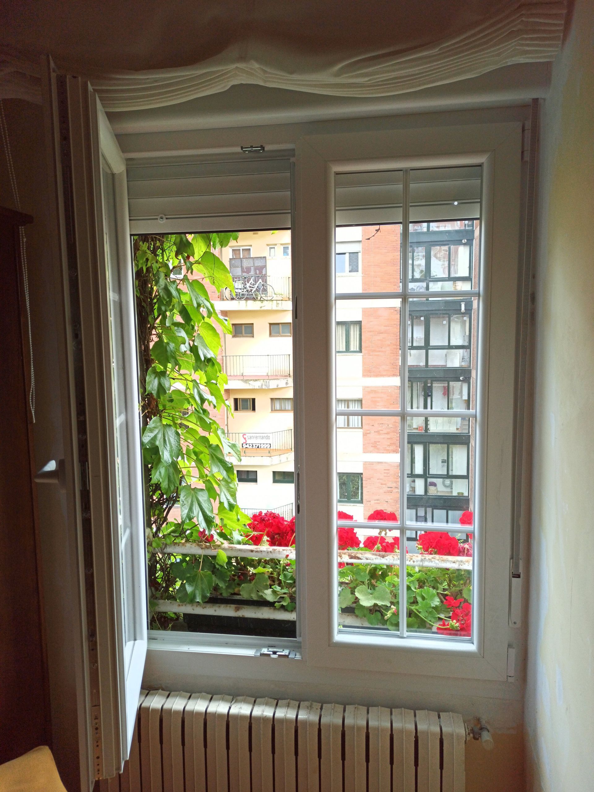 Ventana con marco blanco, con vistas a edificios de apartamentos y vibrantes flores rojas en jardineras, con enredaderas verdes. Un radiador se encuentra debajo.