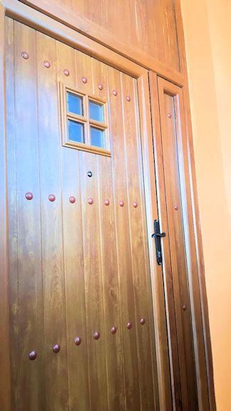 Puerta de madera con una pequeña ventana cuadrada, tachuelas color cobre y manija negra. La puerta está empotrada en una pared naranja claro.