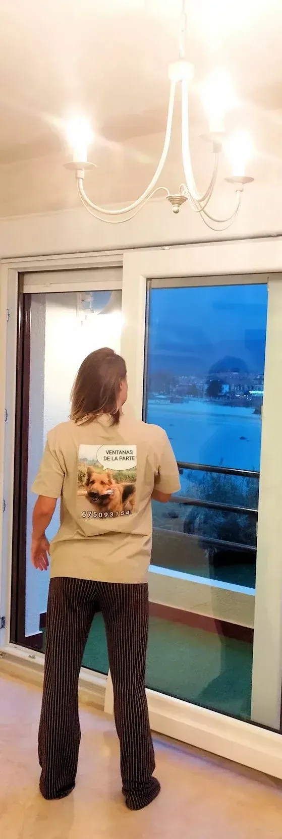 Una persona con cabello largo, vestida con una camisa color canela y pantalones a rayas, mira desde una ventana grande una vista nocturna de un cuerpo de agua.