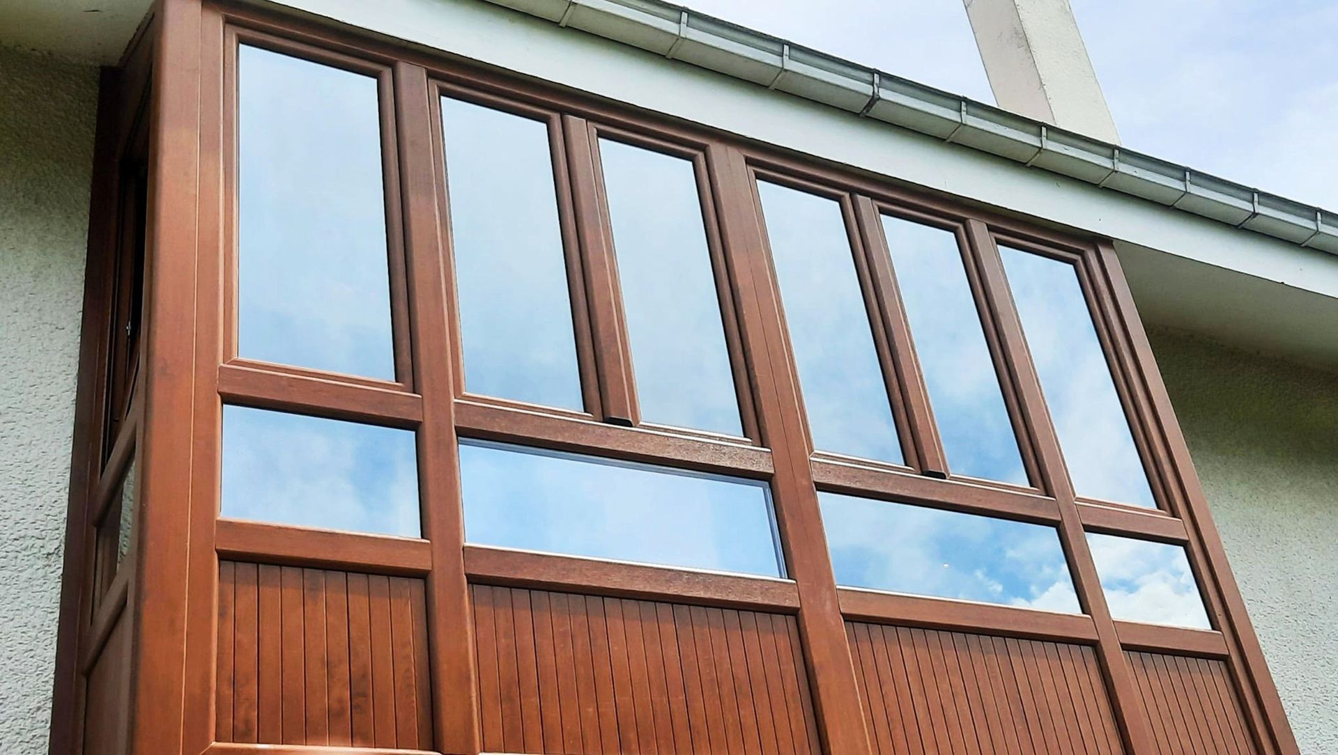 Panel de ventana con marco marrón, múltiples paneles y base de madera decorativa que refleja el cielo. Montado sobre un edificio beige.