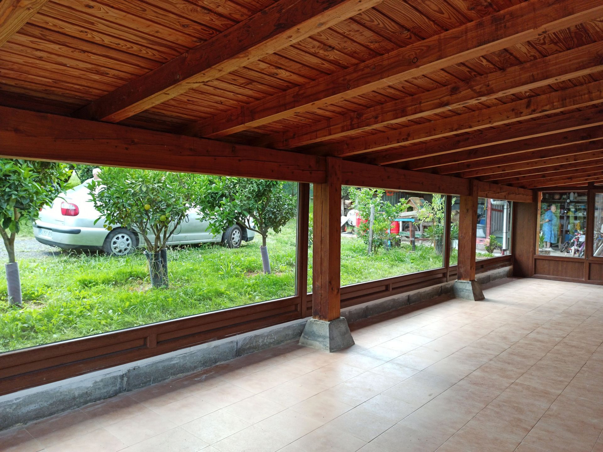 Patio exterior con estructura de madera y suelo de baldosas, abierto a un jardín con un coche y árboles. El techo también es de madera.