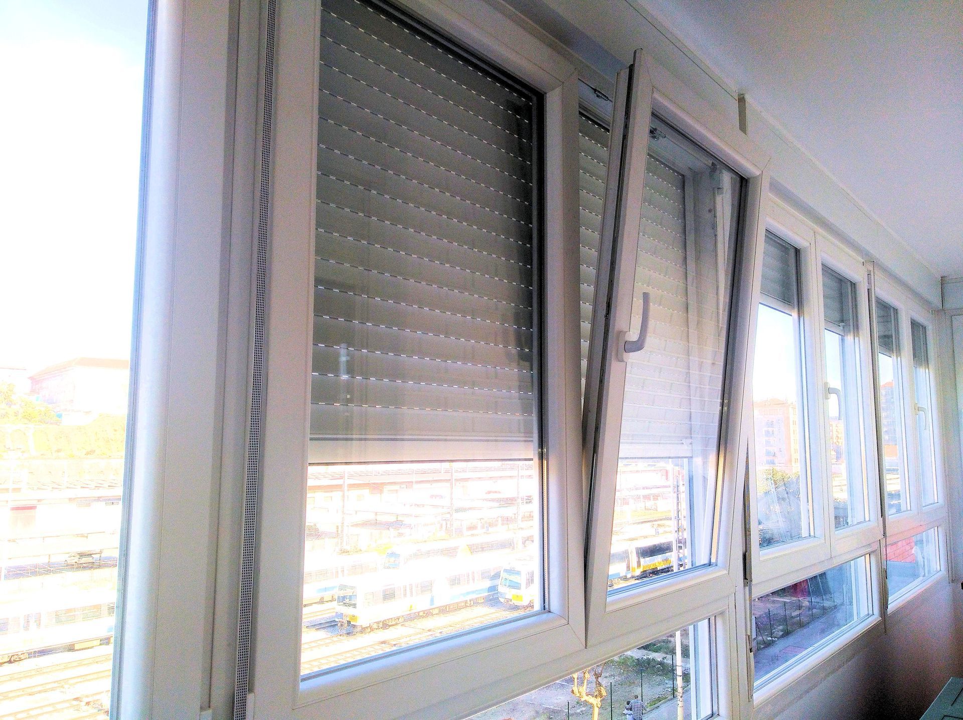Ventana blanca con persiana cerrada y ventana abierta a balcón, con vista a un patio de tren.