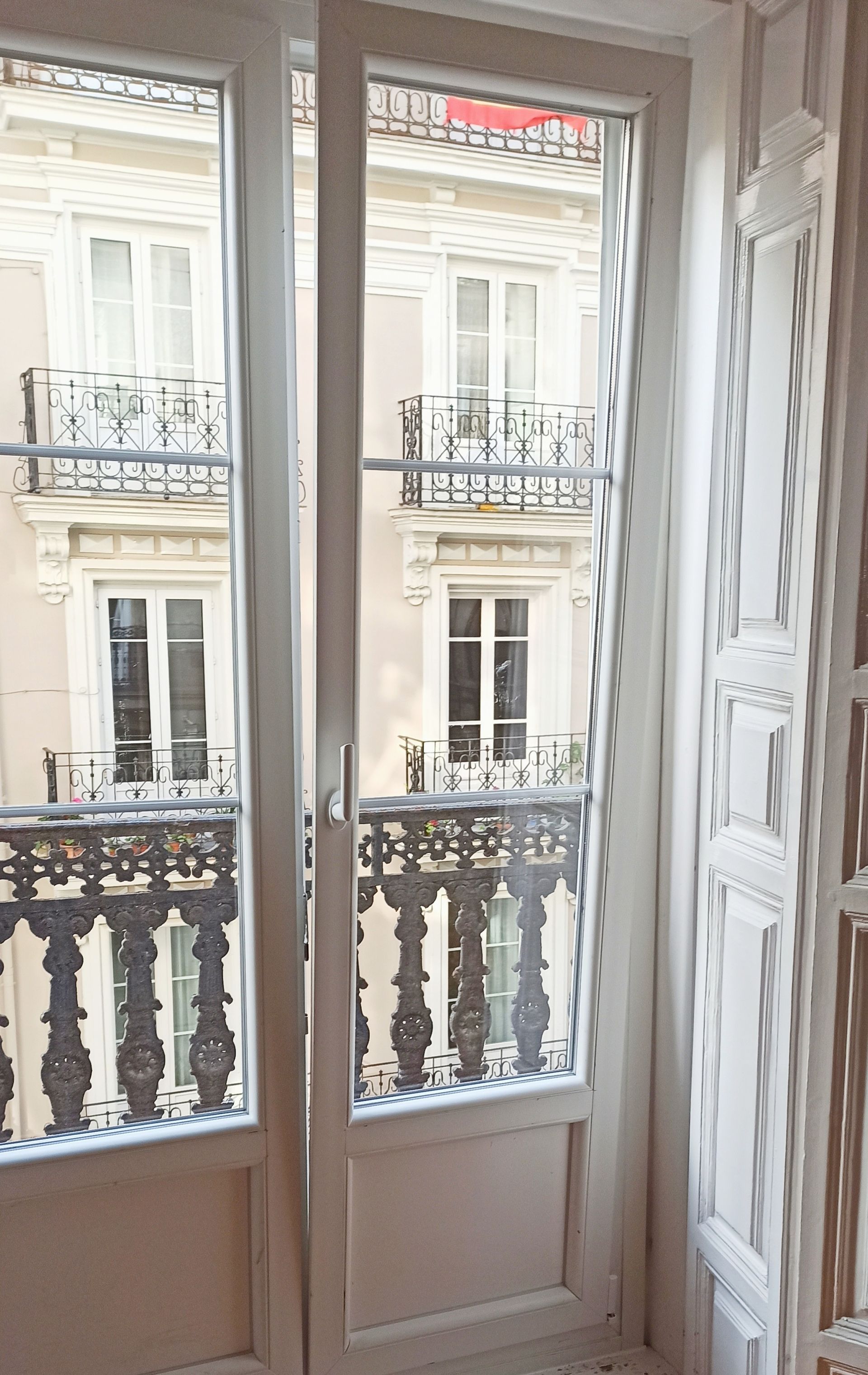 Las puertas francesas blancas se abren a una vista de un edificio con balcones y ventanas adornados; un día soleado.