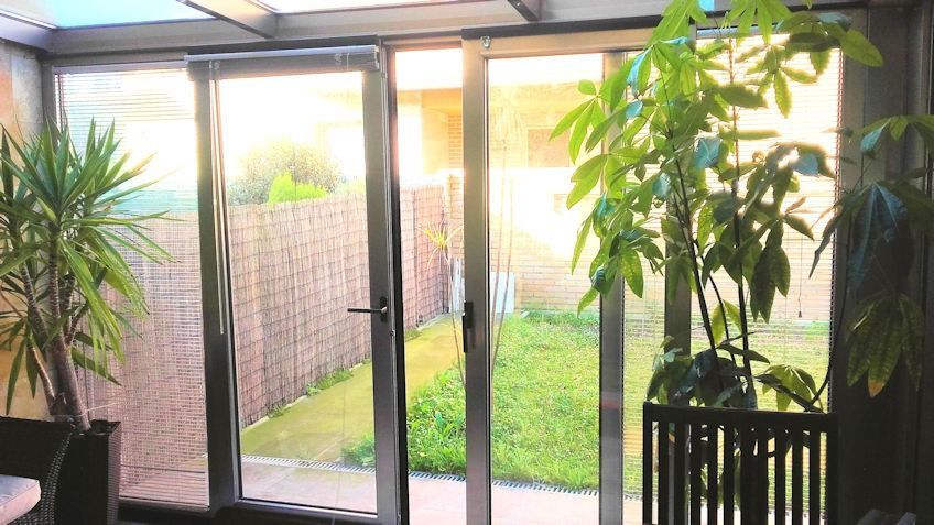 Terraza acristalada con puertas de cristal que dan a un patio trasero con césped y plantas. La luz del sol entra a raudales.