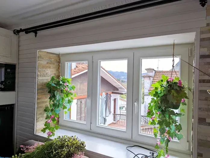Ventana con marcos blancos, flanqueada por paredes de piedra y paneladas, con plantas colgantes y una vista de casas y naturaleza.
