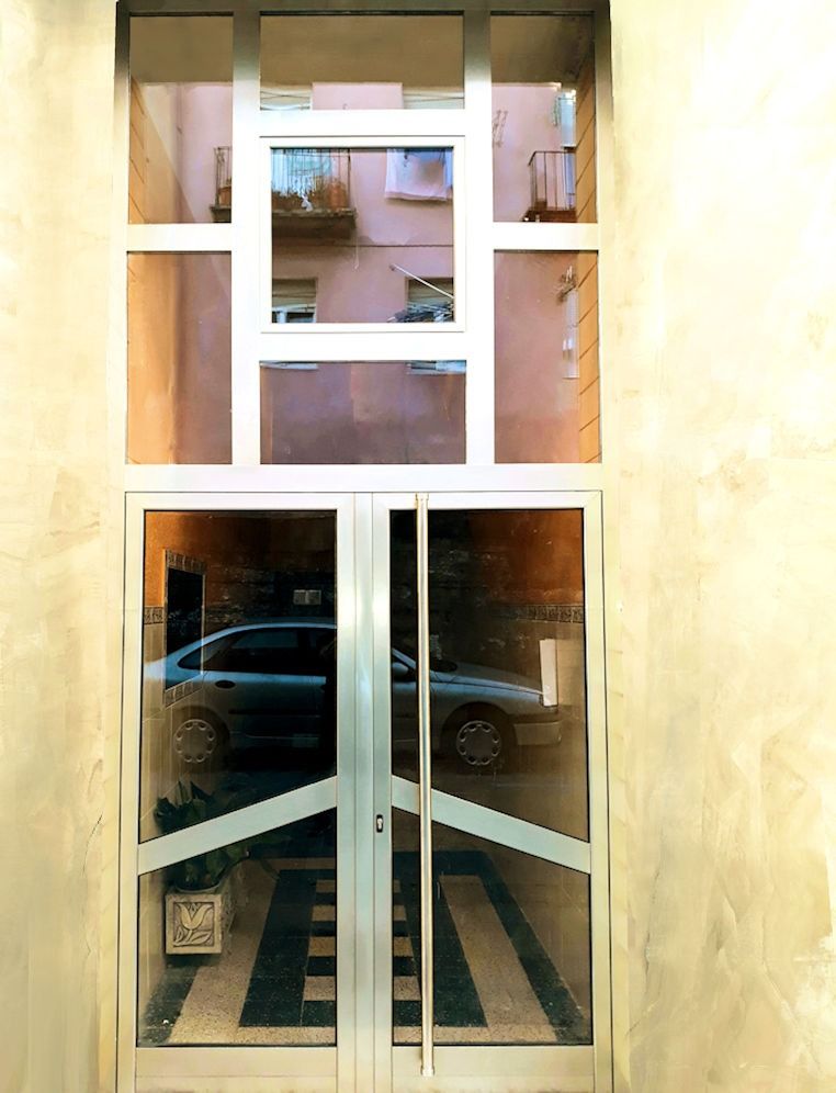 Puertas de cristal con el reflejo de un coche y un patio visible a través de una ventana superior. El edificio tiene paredes de color rosa claro.