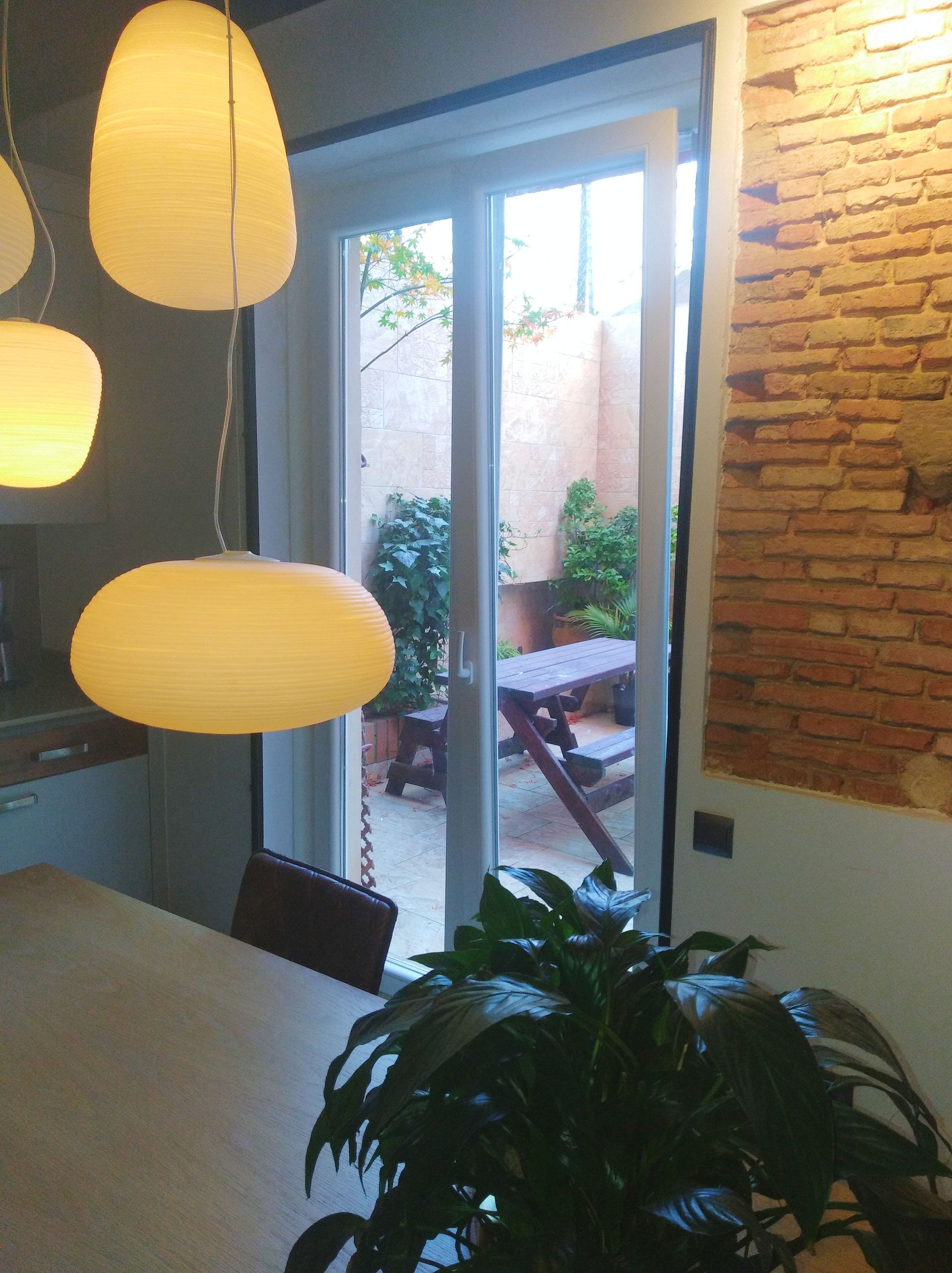 Vista interior con puerta de patio abierta a un pequeño patio; lámparas colgantes en el comedor, pared de ladrillo a la derecha.