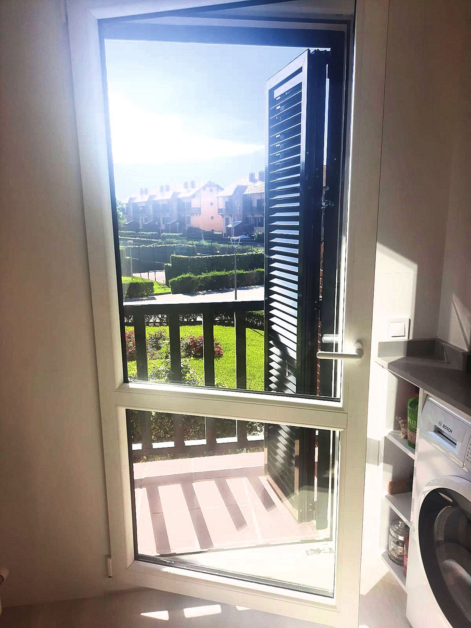 Una puerta da a un balcón soleado con césped verde. La puerta tiene una mosquitera y una contraventana parcialmente abierta.