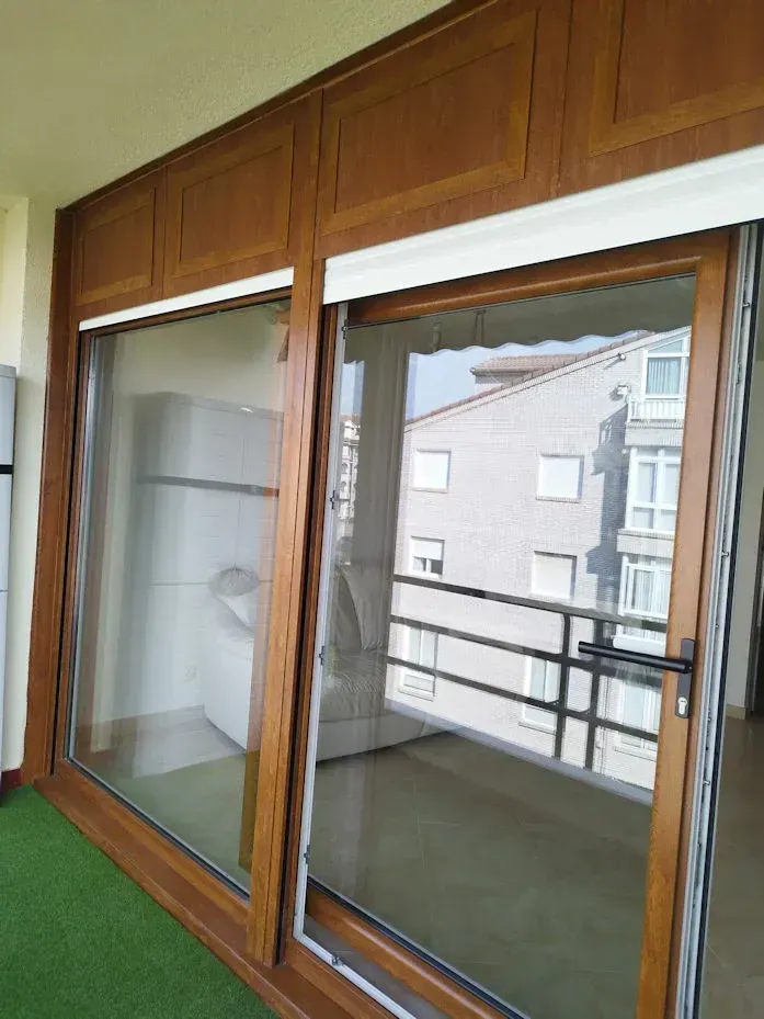 Puerta corrediza de vidrio con marco de madera, con vista a un edificio. El marco es marrón y el interior tiene paredes blancas.