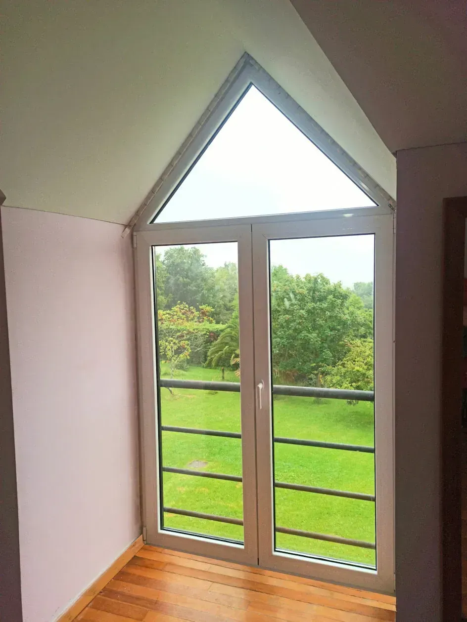Una ventana alta con marco blanco en una habitación con una parte superior triangular tiene vista a un exuberante patio verde con árboles.