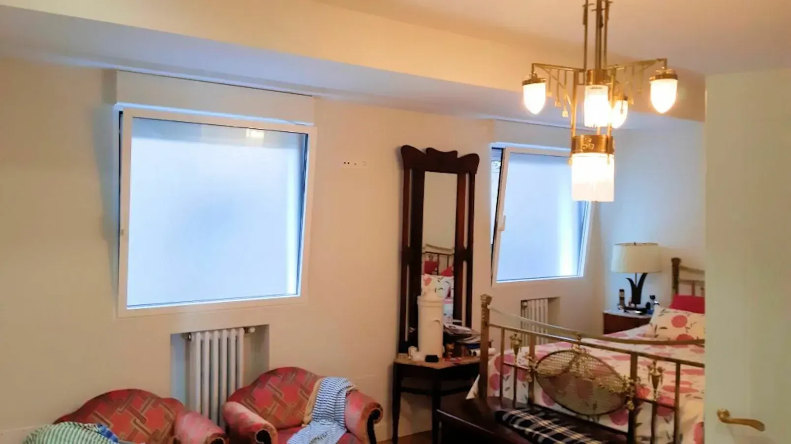 Un dormitorio con dos ventanas, una cama de estilo vintage y una lámpara de araña. También hay sillas, un espejo de cuerpo entero y un radiador.