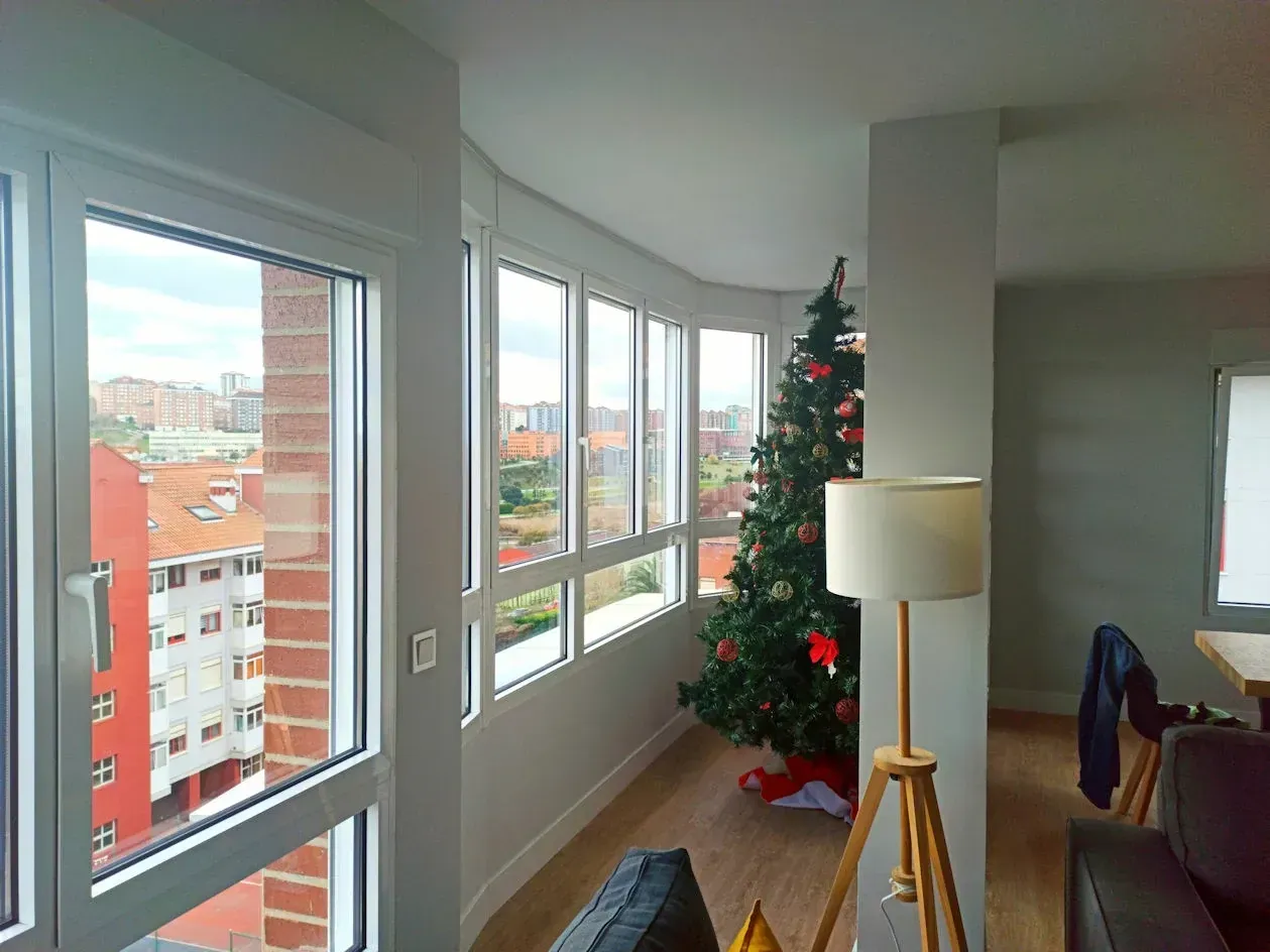 Vista interior de una habitación con grandes ventanales que ofrecen vistas a la ciudad. Un árbol de Navidad se alza junto a las ventanas; también hay una lámpara y muebles.