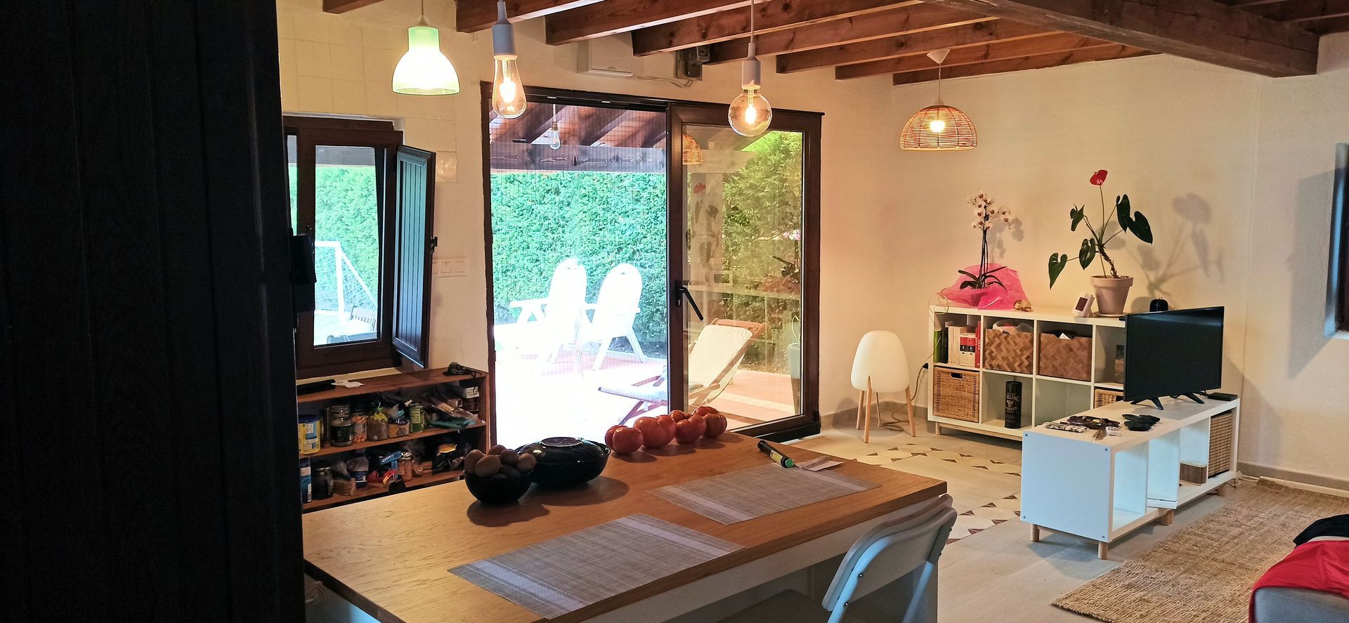 Vista interior de un luminoso salón con techo de vigas de madera y vistas a un patio exterior. Hay una isla de cocina y un televisor.