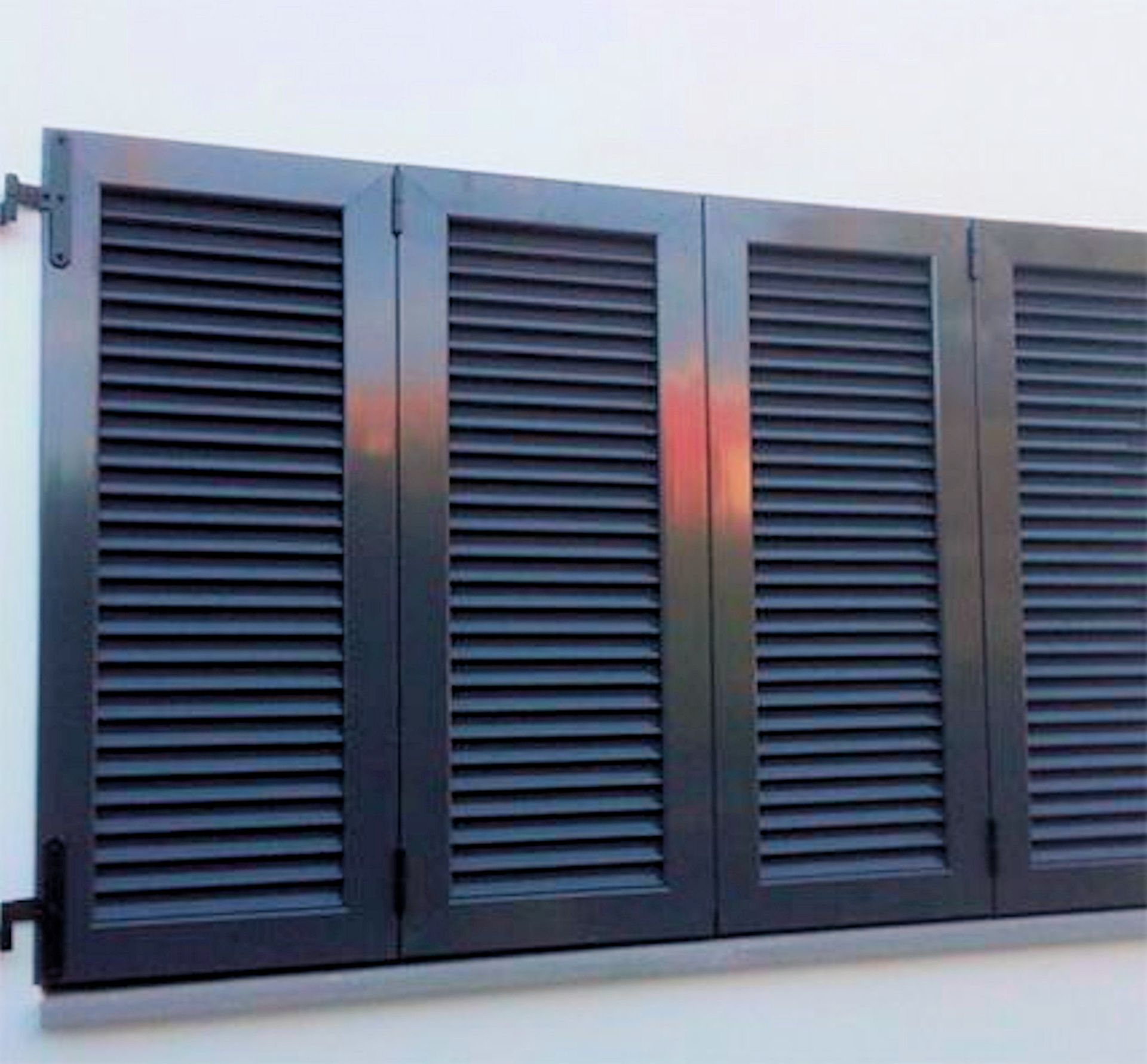 Cuatro contraventanas de color gris oscuro unidas a una pared blanca, parcialmente abiertas.