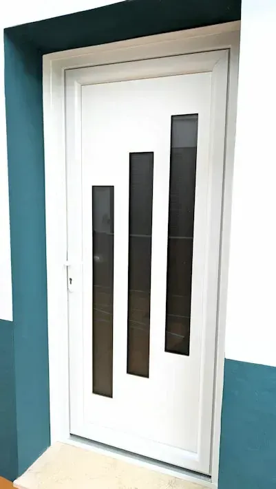 Puerta blanca con tres rectángulos verticales oscuros. La puerta está situada contra una pared blanca y verde azulado.