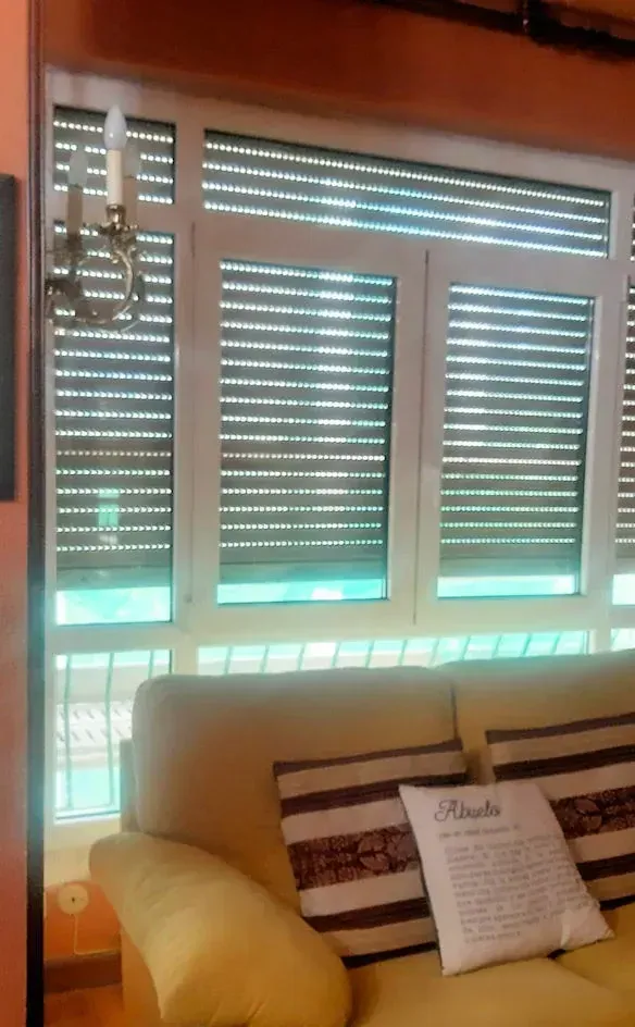 Una ventana con persianas marrones cerradas sobre un sofá beige con cojines. Un candelabro decorativo está colgado en la pared.