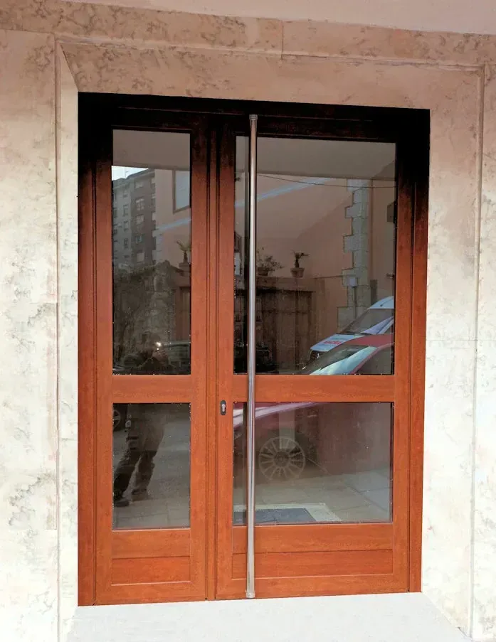 Puertas dobles de madera con paneles de vidrio colocados en un marco de mármol; una persona se refleja en el vidrio.
