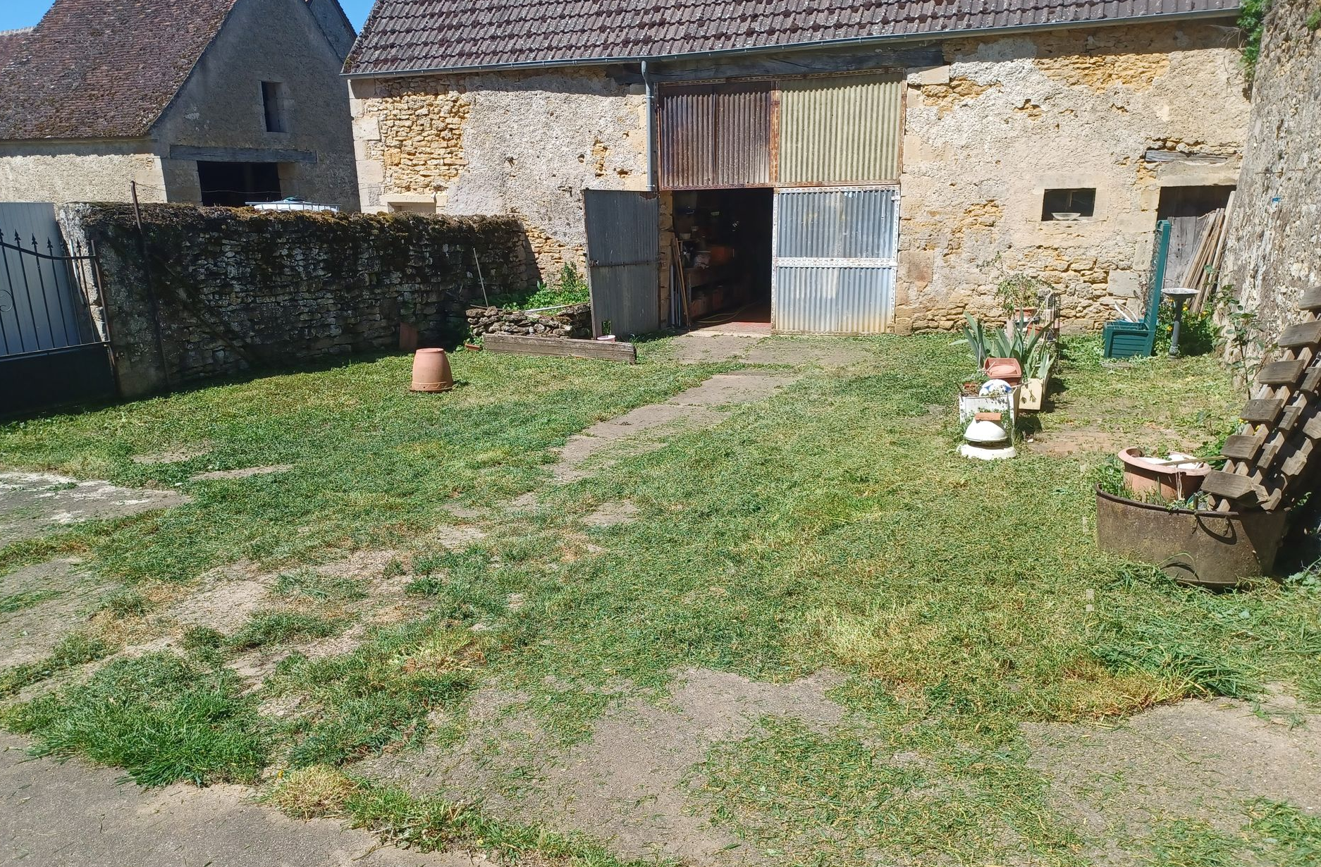 Cour de ferme en pierre avec pelouse, chemin de gravier, pots de fleurs et haie sous un soleil éclatant