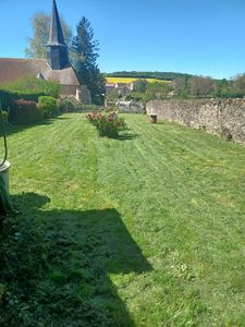 Jardin ensoleillé avec pelouse verte, mur de pierre et clocher d'église en arrière-plan