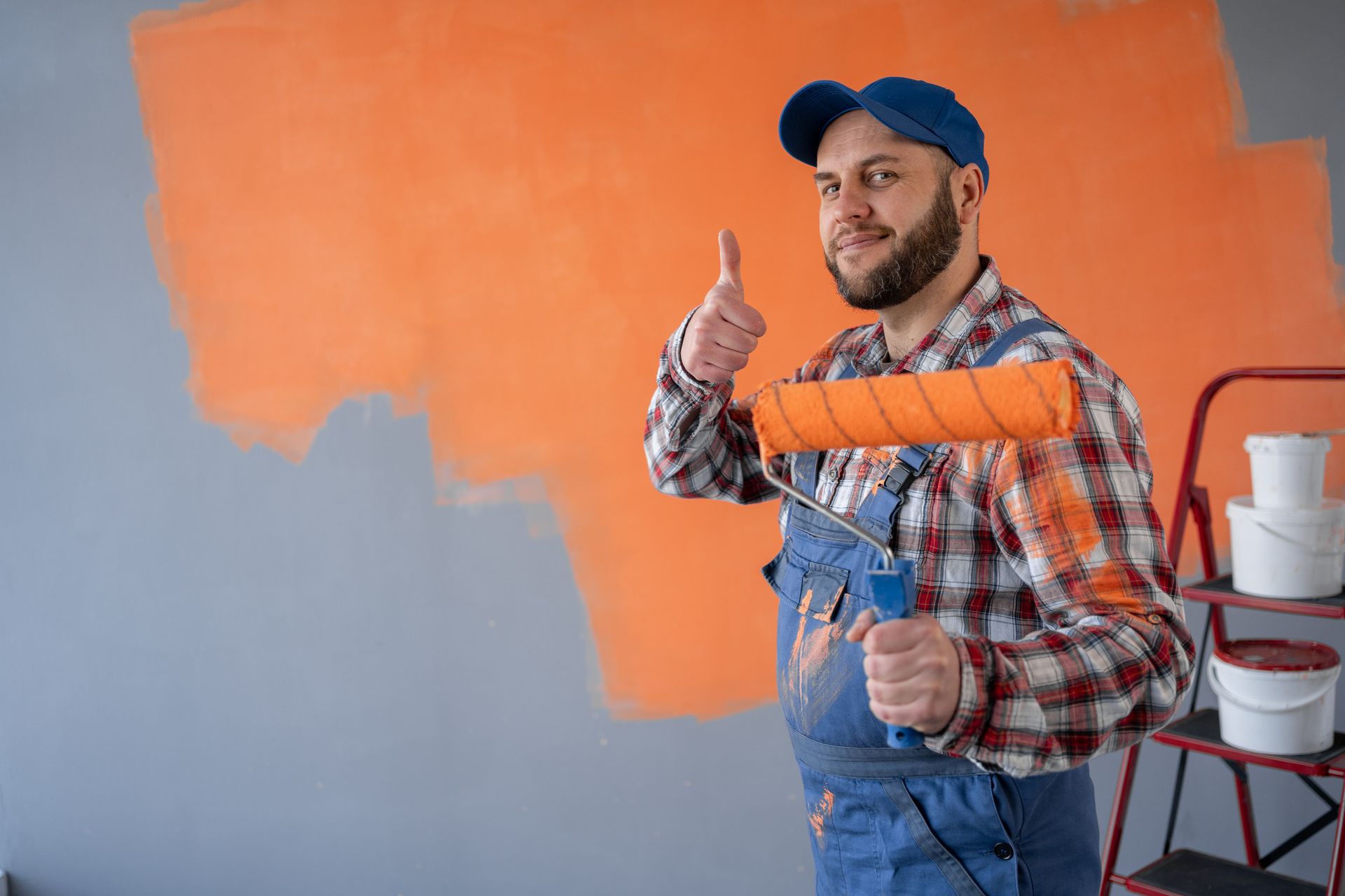 Un pintor con mono y gorra azules hace un gesto de aprobación con el pulgar mientras sostiene un rodillo con pintura naranja en la pared.