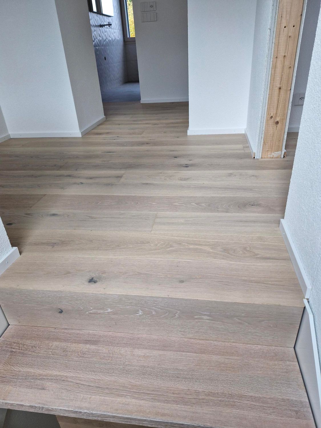 Un parquet clair recouvre un escalier intérieur et un couloir, avec des murs blancs et un pilier en bois brut.