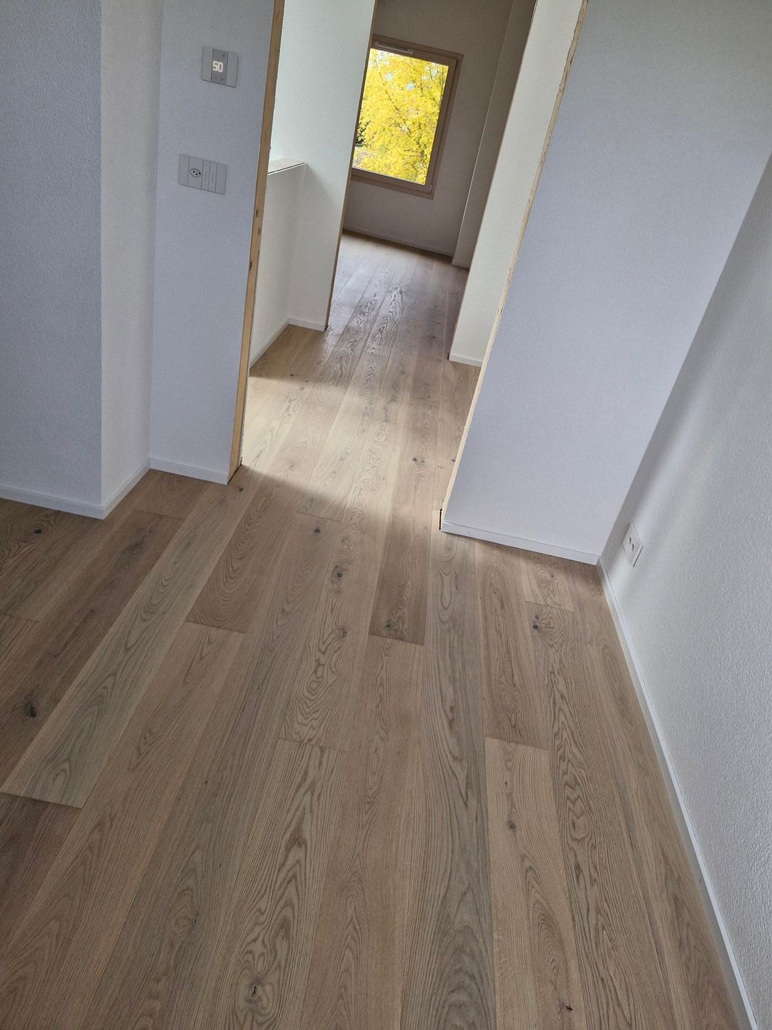 Un couloir en bois mène à une porte ouverte offrant une vue sur un feuillage jaune vif à travers une fenêtre.