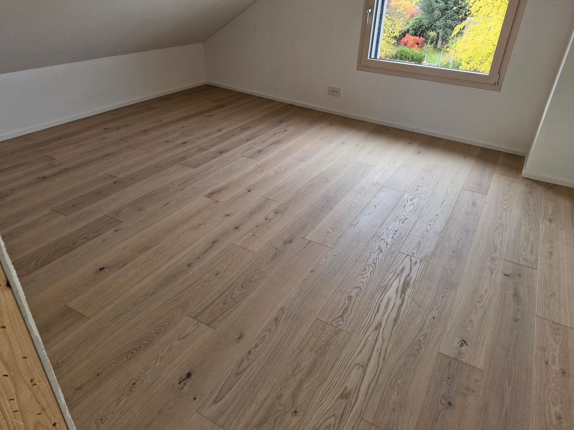 Une chambre mansardée lumineuse avec un parquet en bois clair et une fenêtre donnant sur la verdure.