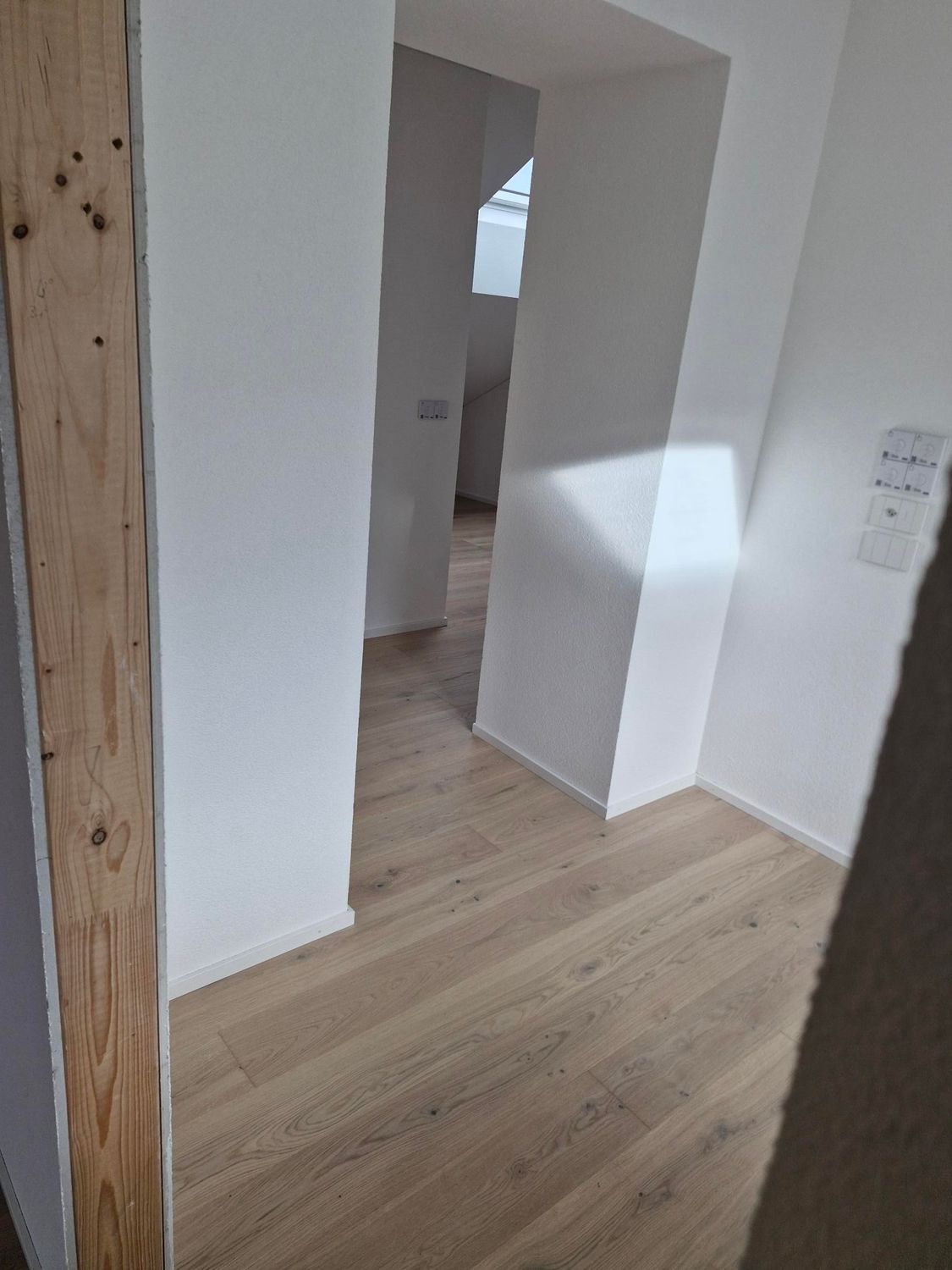 Vue à travers une porte donnant sur un couloir au parquet clair et aux murs blancs unis, baigné d'une lumière naturelle vive.