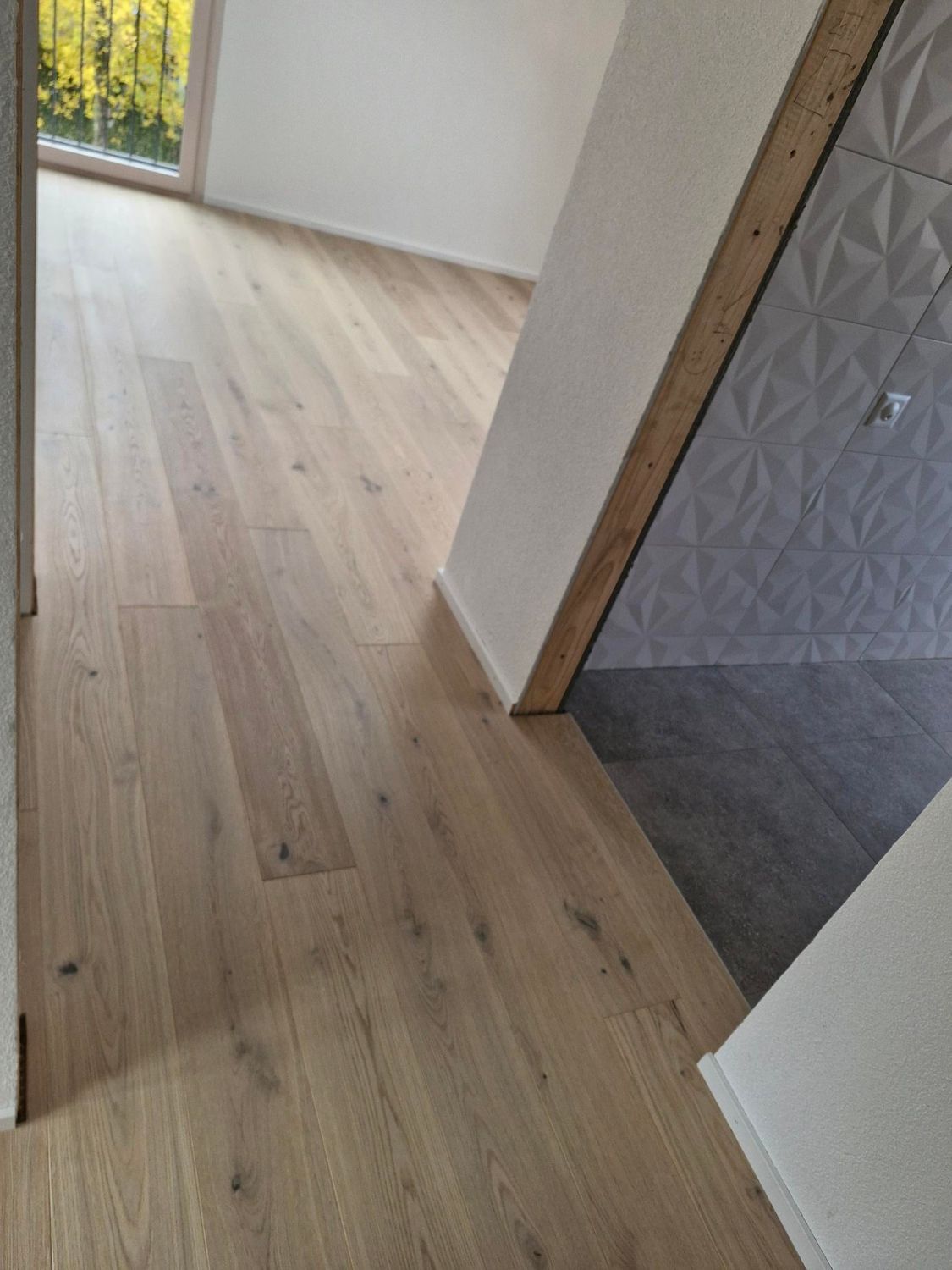 Un couloir au parquet en chêne clair débouche sur une zone carrelée de carreaux muraux géométriques gris.