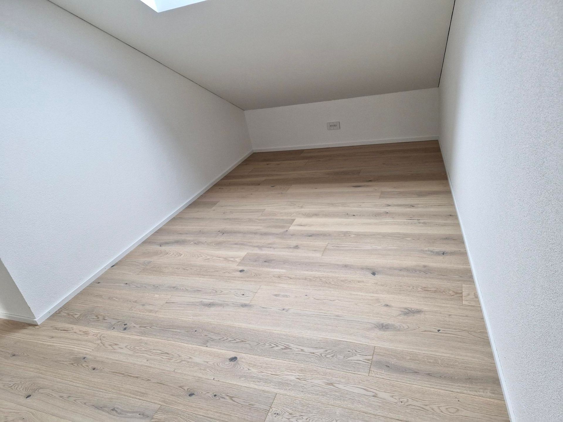 Une pièce mansardée vide, avec des murs blancs en pente, un parquet clair et une lucarne rectangulaire au-dessus.