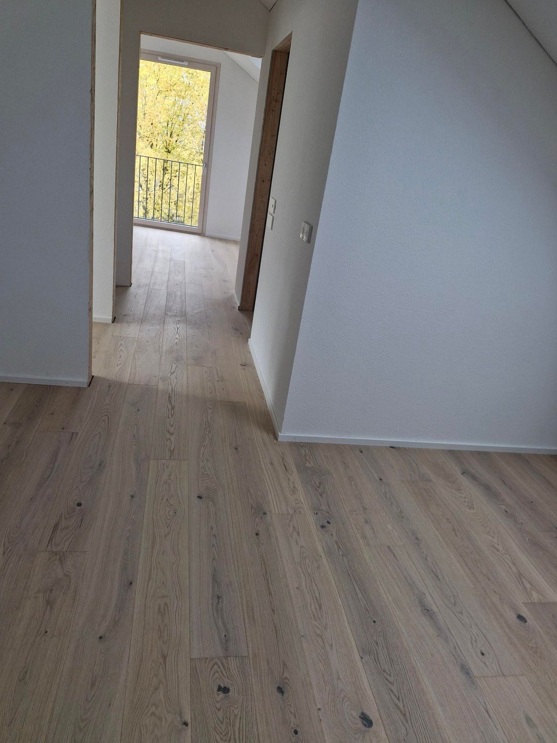 Un couloir lumineux avec un parquet clair mène à une porte qui s'ouvre sur un balcon avec des arbres visibles à l'extérieur.