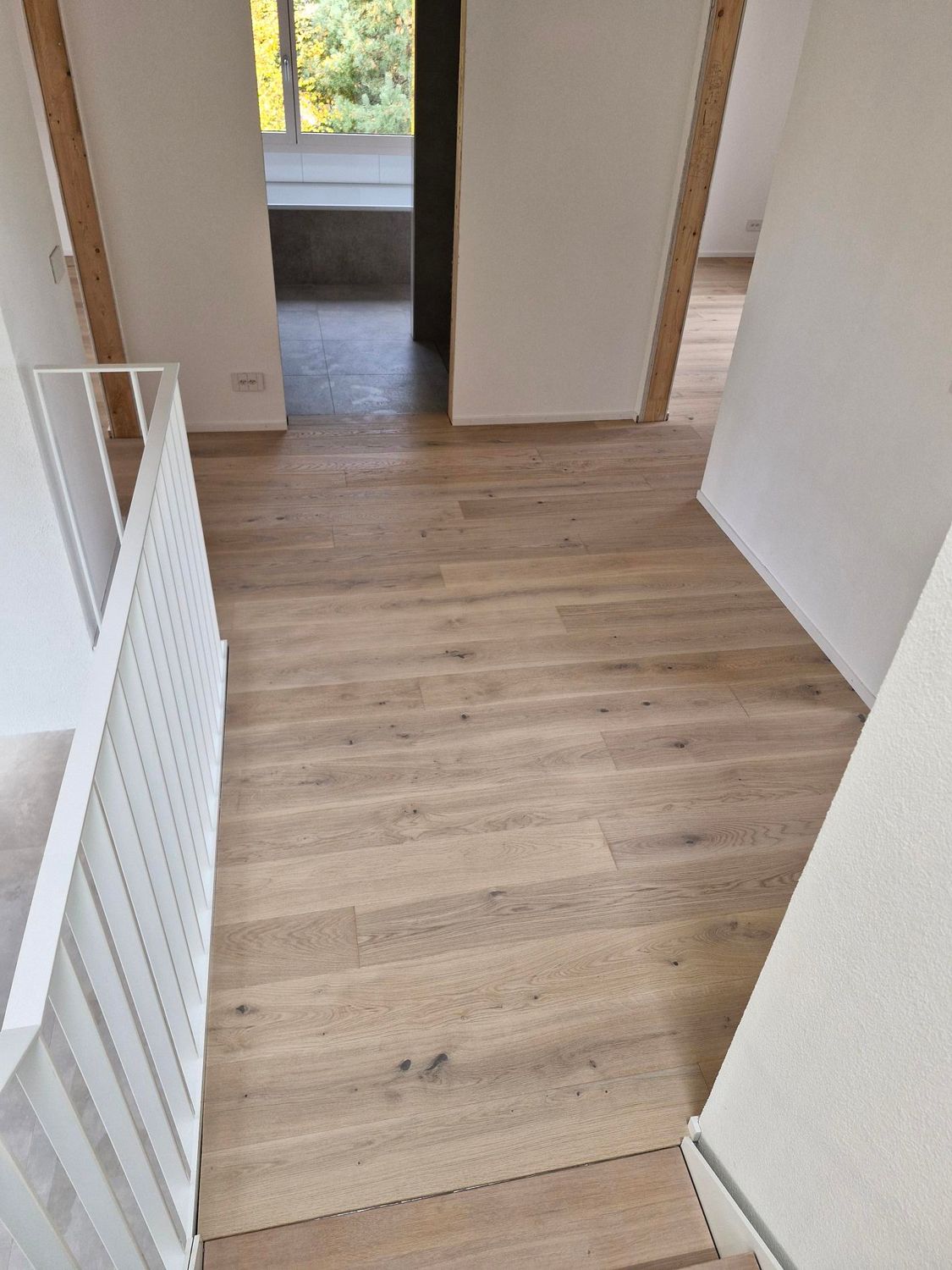 Vue de dessus d'un couloir au sol en bois clair menant à une porte ouverte avec une rampe blanche sur la gauche.