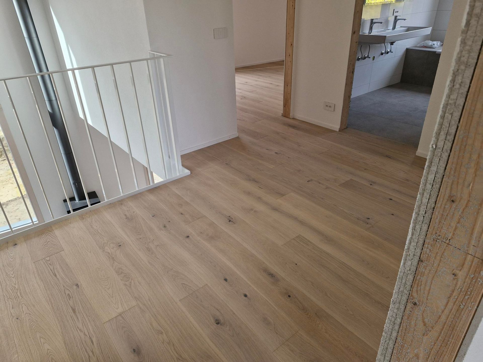 Vue intérieure d'un couloir avec parquet clair, rampe en métal blanc et vue sur une salle de bains.