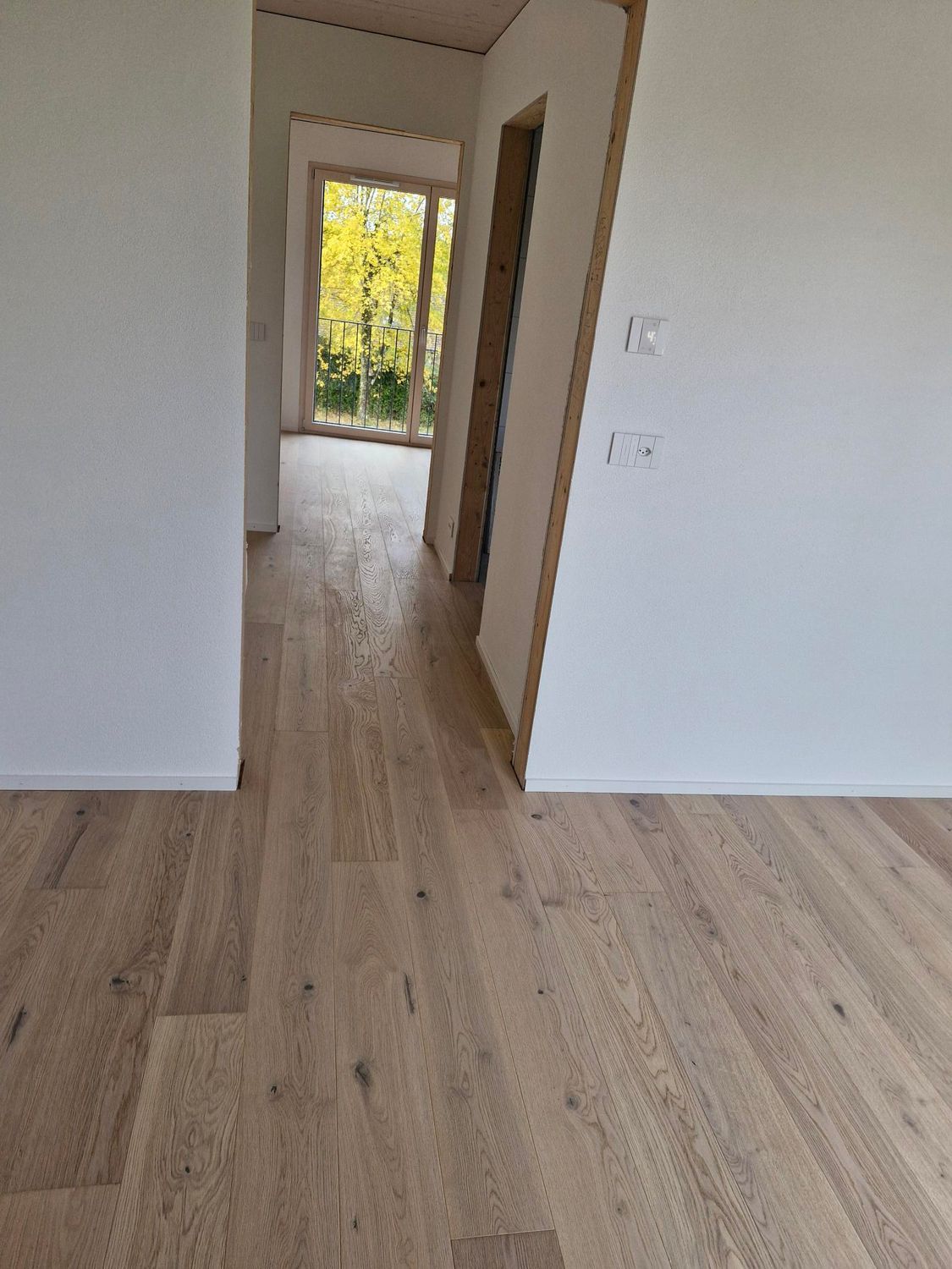 Vue intérieure montrant un parquet clair se prolongeant d'une pièce ouverte à travers une porte jusqu'à un couloir.