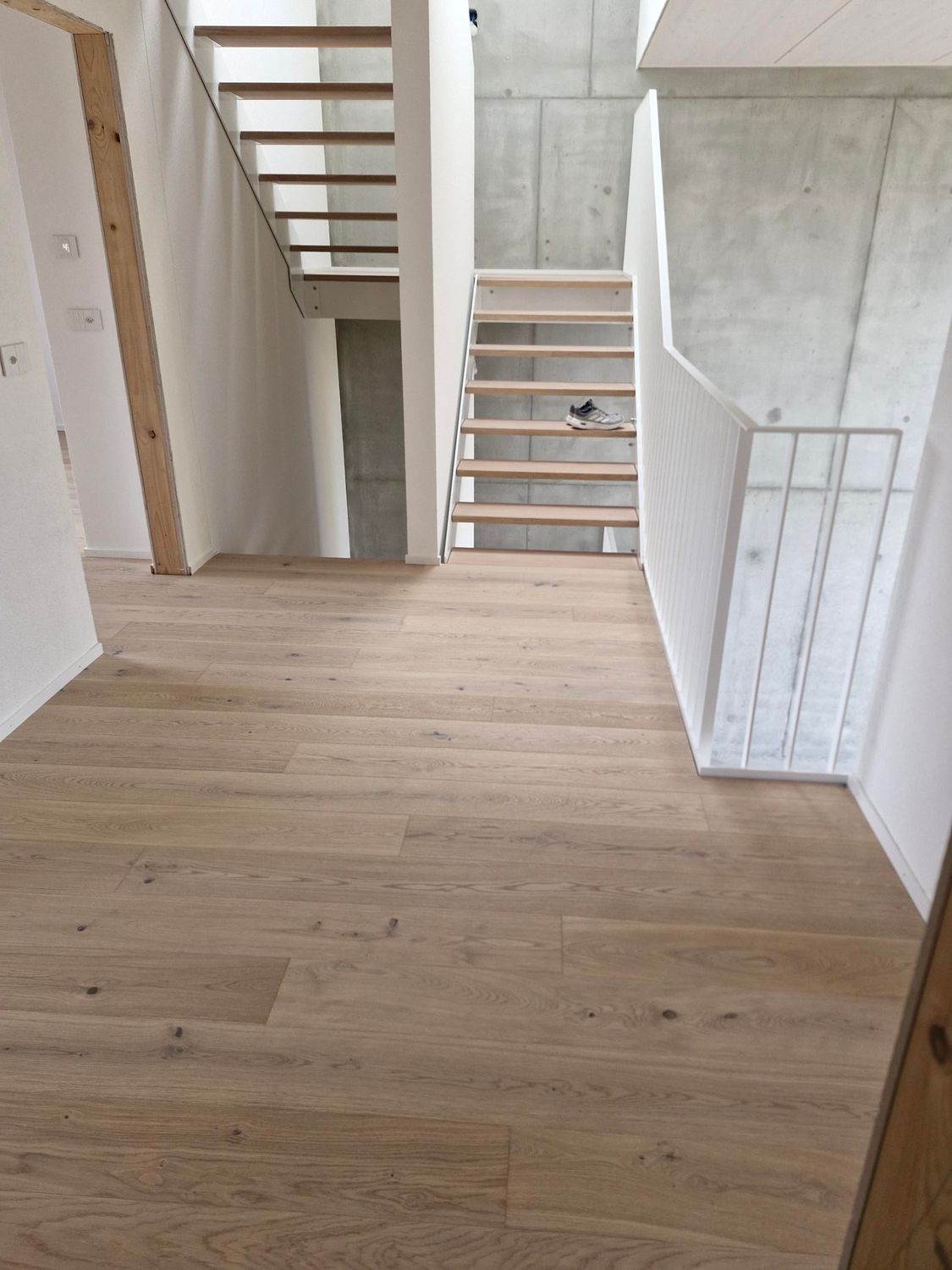 Un palier intérieur minimaliste avec parquet clair, un mur en béton et deux escaliers en bois à marches ouvertes.
