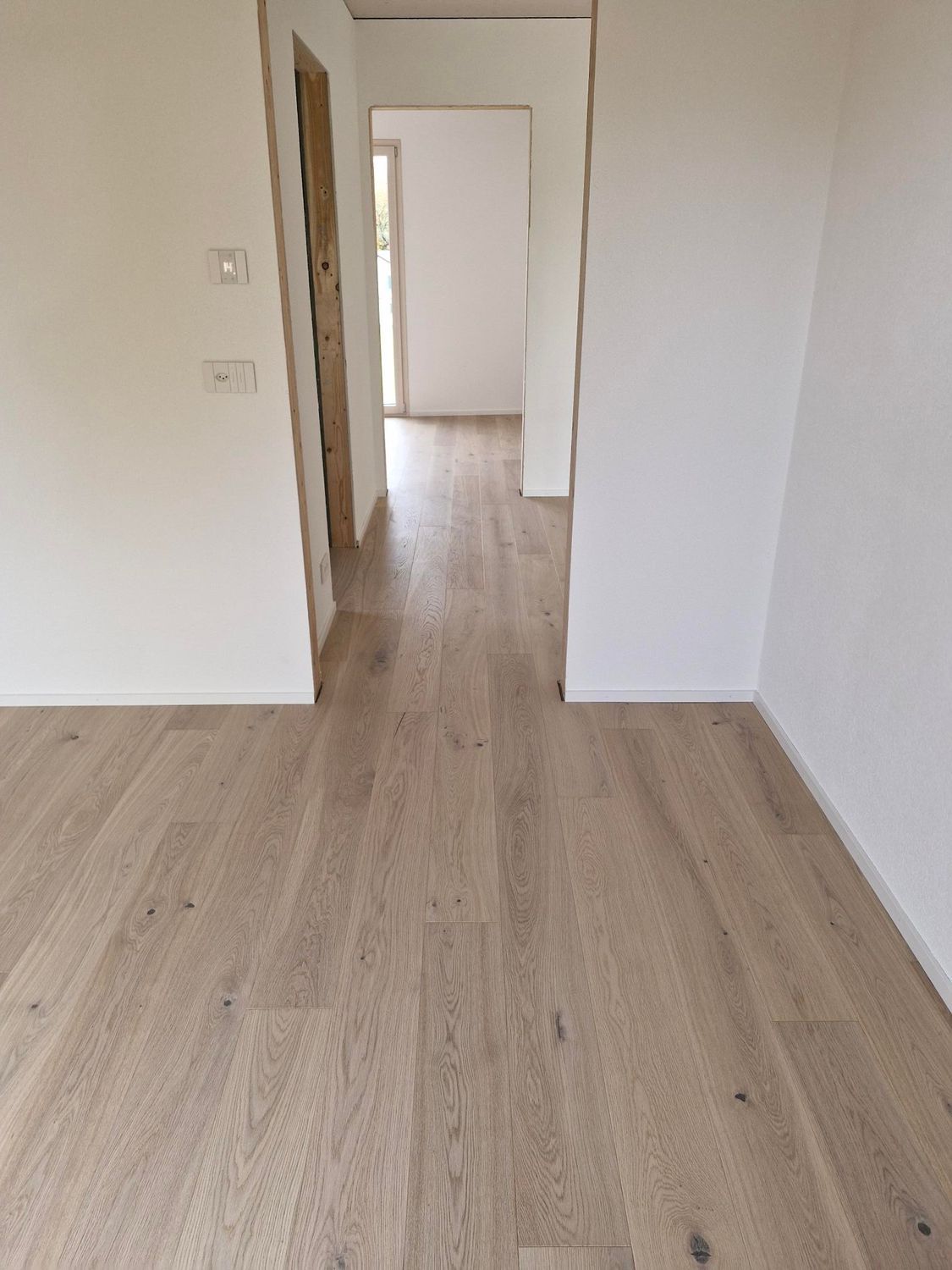 Vue intérieure lumineuse et minimaliste d'un parquet clair menant, par une porte ouverte, à un couloir aux murs blancs.