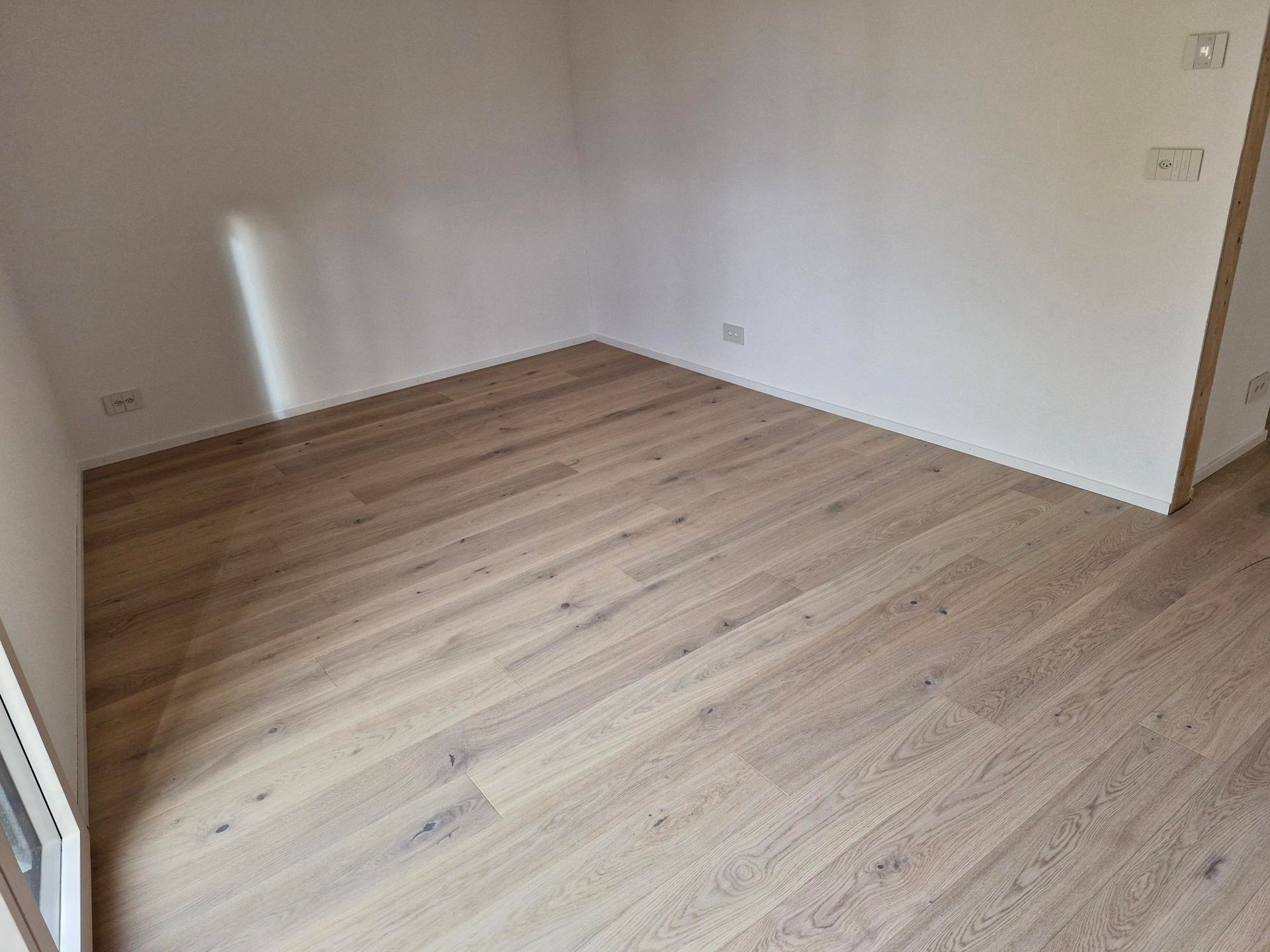Une pièce vide avec un parquet clair, des murs blancs et des prises électriques.