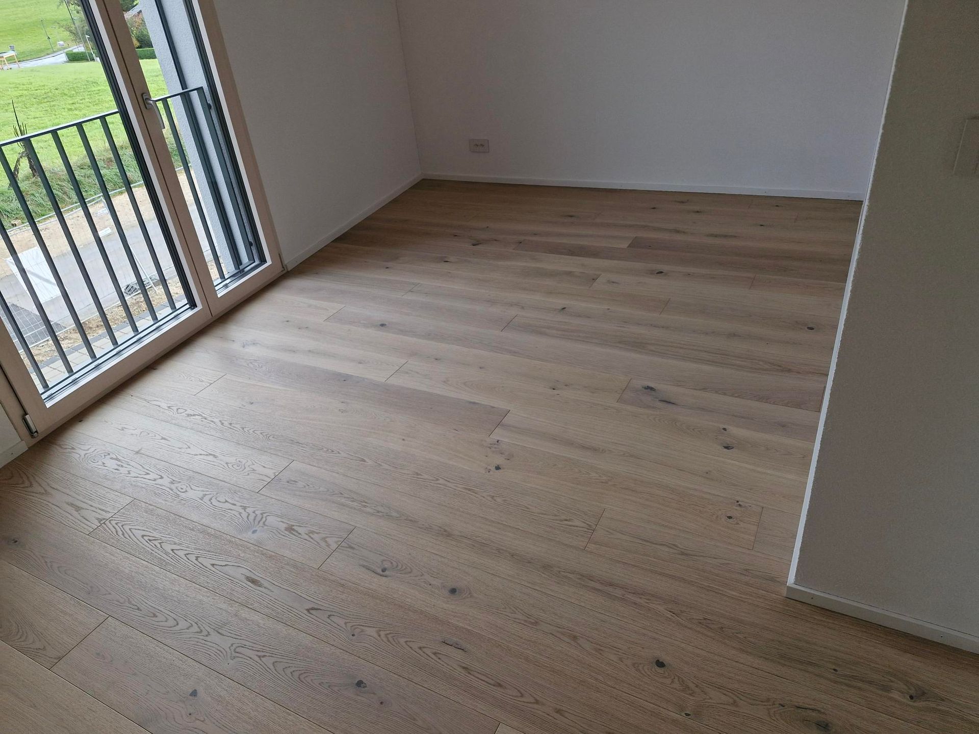 Parquet clair dans une pièce vide avec une grande porte vitrée donnant sur un balcon extérieur.