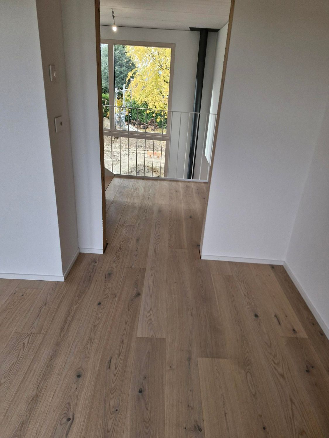 Vue à travers une porte sur une autre pièce dotée d'un parquet clair et d'une grande fenêtre donnant sur des arbres.