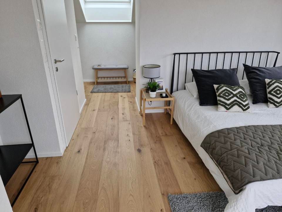 Une chambre minimaliste avec parquet clair, un lit aux oreillers noirs et une petite table de chevet sous une lucarne.