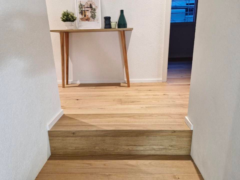 Une marche en bois mène à un couloir doté d'un parquet clair, de murs blancs et d'une console en bois décorée.