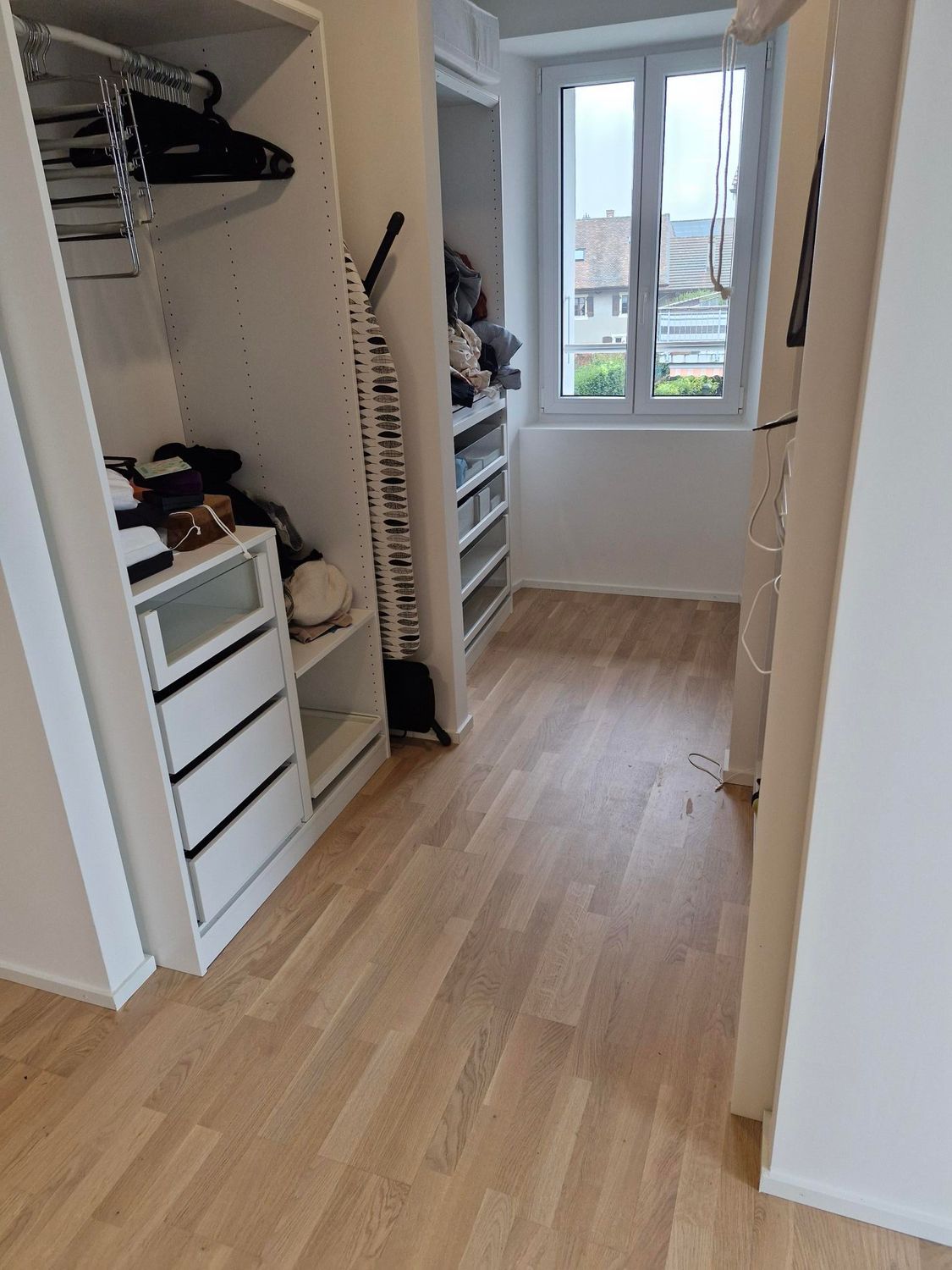 Un petit dressing avec parquet clair, comprenant une commode blanche, des rangements suspendus et une fenêtre lumineuse.