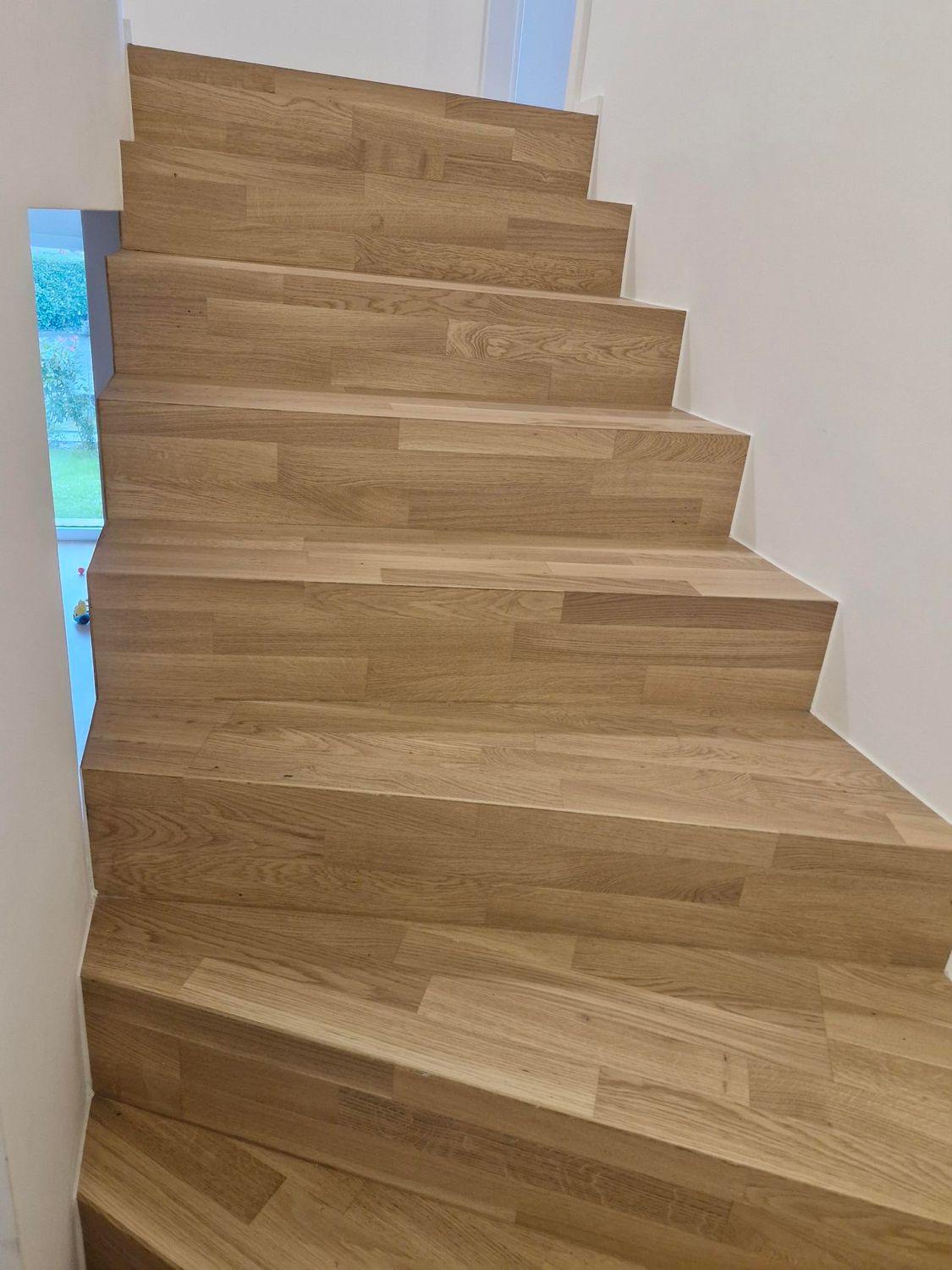 Un escalier aux marches en bois clair dont le grain est visible, situé entre des murs blancs.