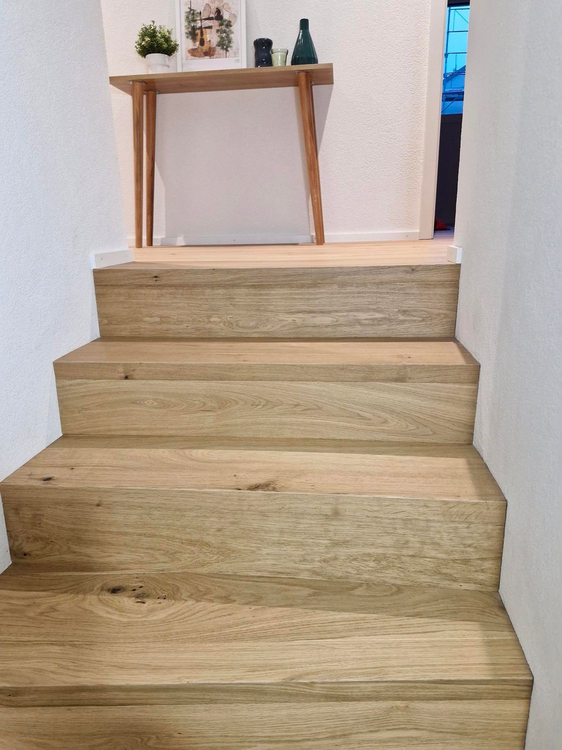 Un escalier en bois clair monte entre deux murs blancs unis, avec une fenêtre visible sur la gauche.