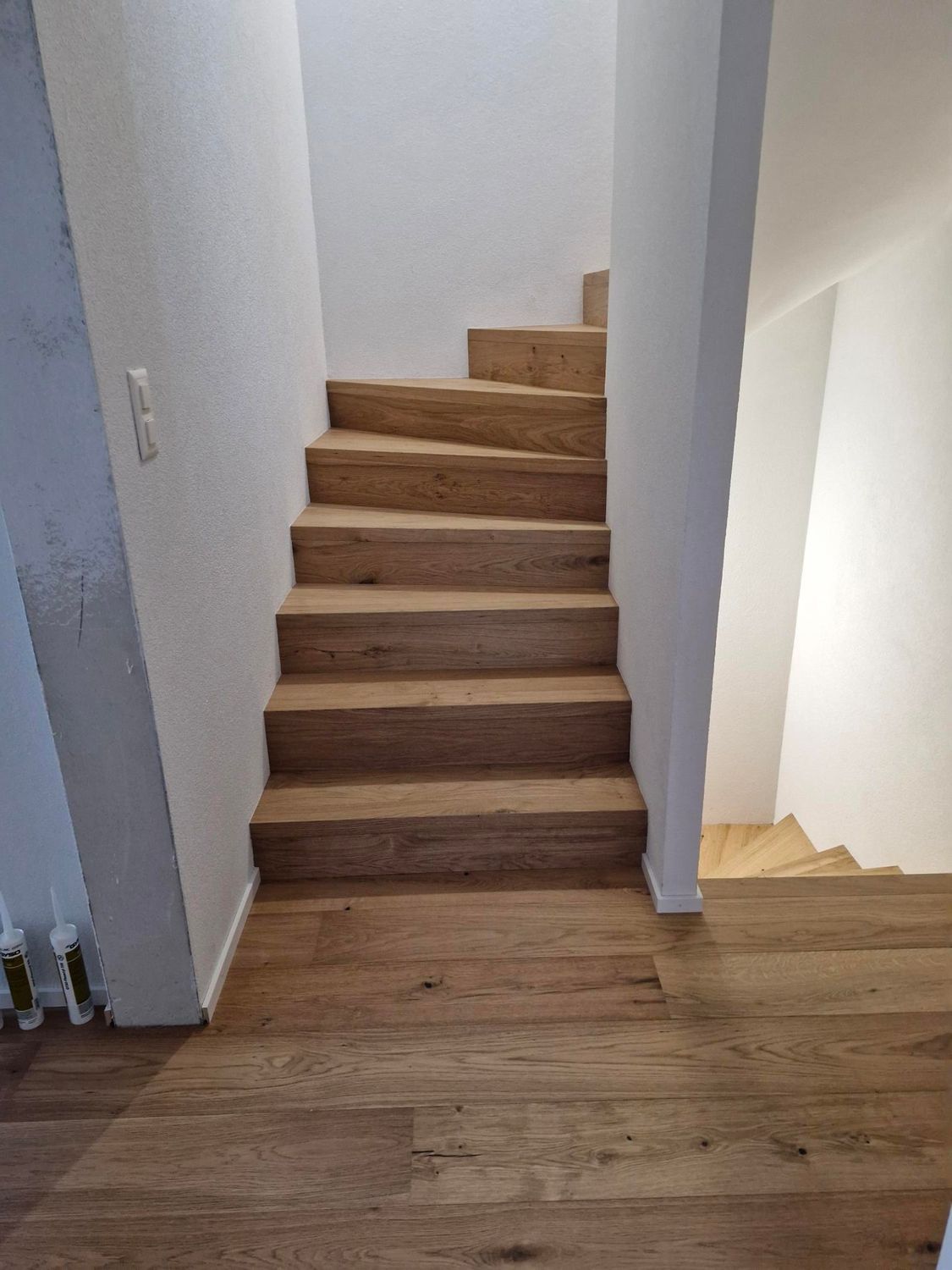 Un escalier en bois à plusieurs marches monte depuis un palier au sol également en bois, entre deux murs blancs unis.