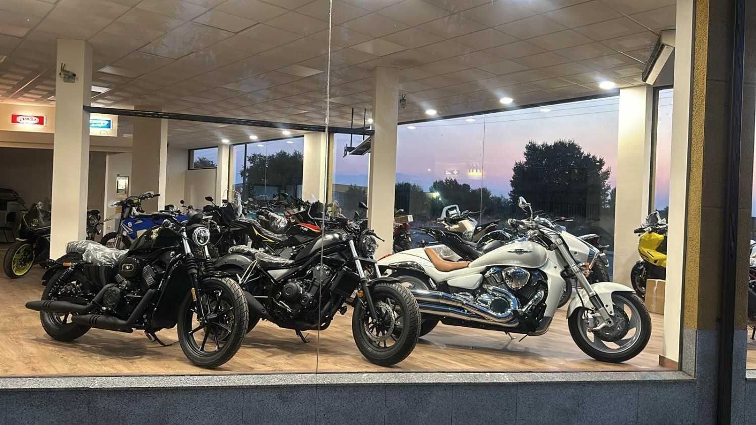 Sala de exposición de motocicletas con una motocicleta todoterreno blanca y gris en exhibición y un scooter en primer plano.